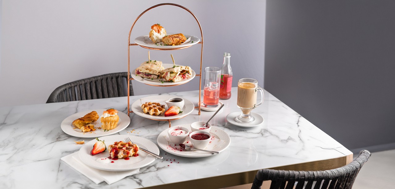 Tiered afternoon-tea stand displays sandwiches and pastries, accompanied by waffles, jam, sliced strawberries, a latte, and pink soda. Set on a marble table in a minimalist café with wicker chairs.